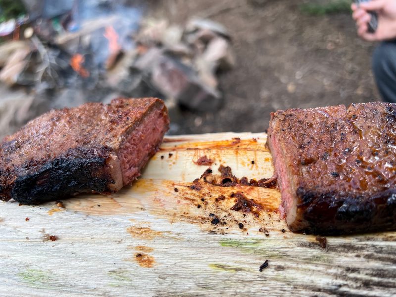 Two seared steaks resting on a split log beside a campfire
