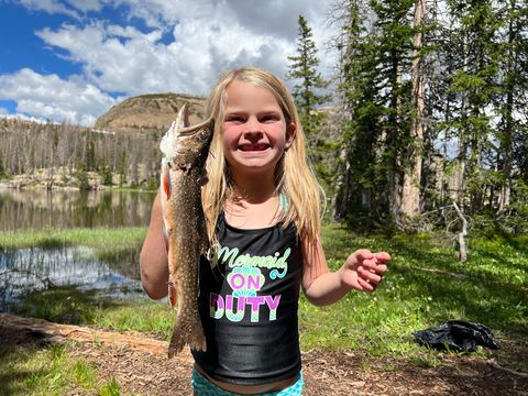 A smiling girl holding up a trout in a grassy meadow beside a mountain lake