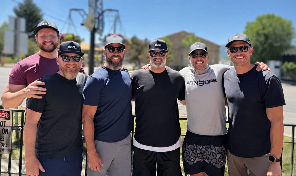 Six Dad Camp dads in matching dad. hats and sunglasses, arms around each other, smiling on a sunny day