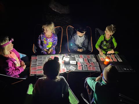 Kids in camp chairs playing cards at night around a table lit by headlamps and a small lantern