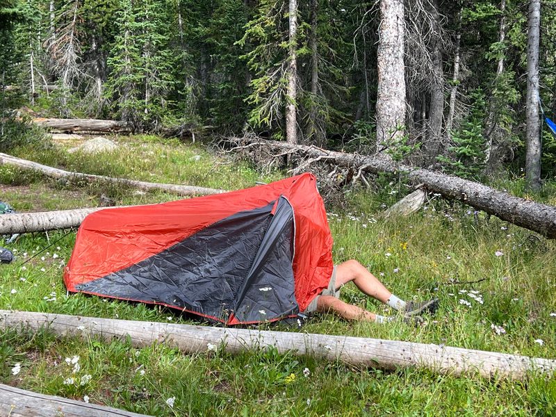 Someone lying half inside a small red tent in a grassy meadow, legs and boots sticking out the door, among fallen logs and pines