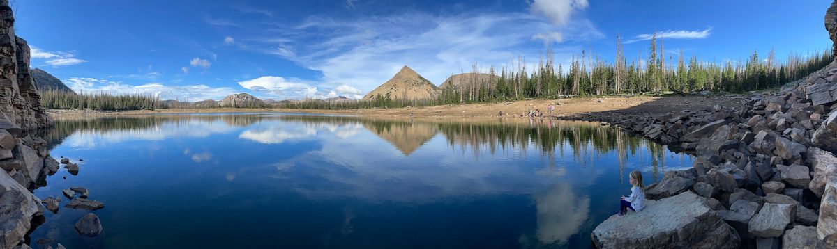 Panorama of a still mountain lake reflecting a pointed peak, pines and clouds, with a child sitting on the boulders at the right shore