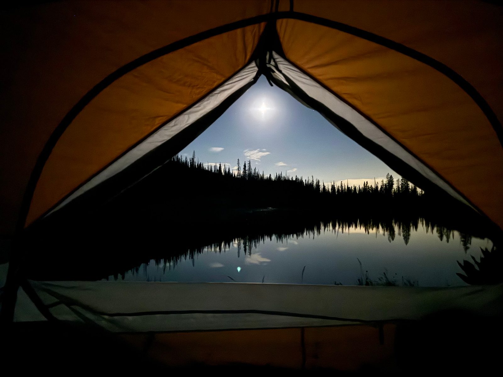 Night view through an open tent door: a bright moon over a still lake lined with dark pines and their reflections