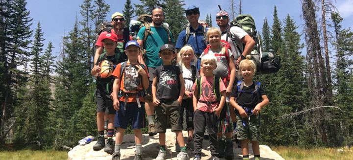 Dad Camp 2018 group photo: dads and kids with backpacks standing together on a big rock among pine trees, Wall Lake trip