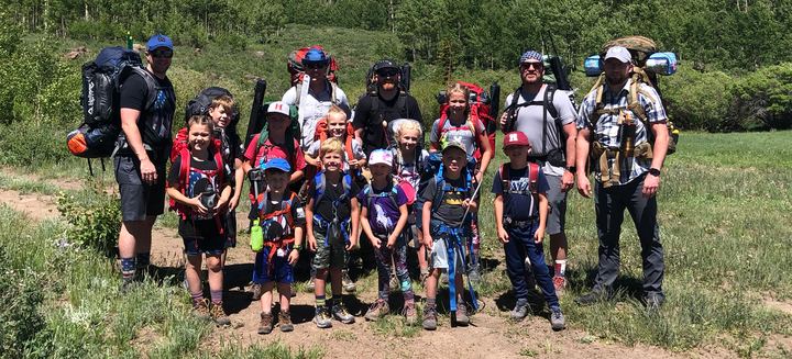 Dad Camp 2019 group photo: dads and kids with backpacks on a trail through a green meadow, Deer Creek Lake trip