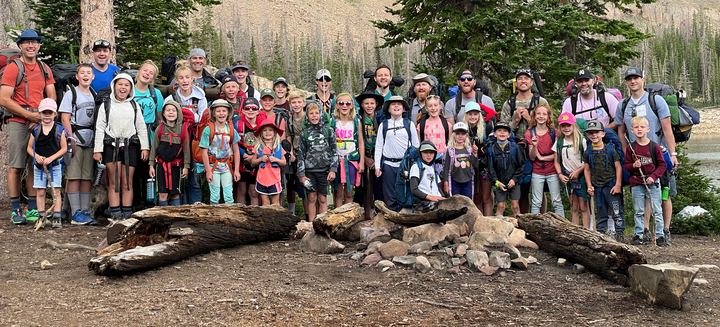 Dad Camp 2021 group photo: dads and kids with backpacks at camp, behind a stone fire ring and logs, with pines and cliffs behind