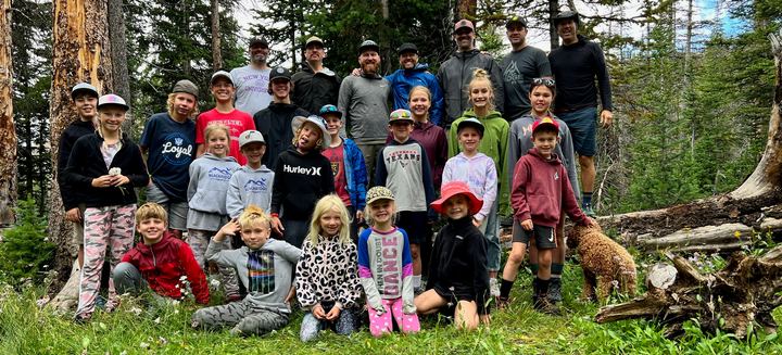 Dad Camp 2022 group photo: dads and kids gathered among pine trees in the woods, some kids sitting in the grass, with a doodle dog, Haystack Lake trip