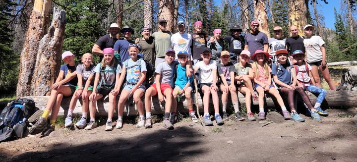 Dad Camp 2024 group photo: kids sitting on a big log with dads standing behind them among tall pines, Grandaddy Lake trip
