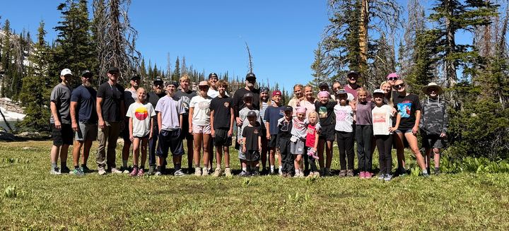Dad Camp 2026 group photo: dads and kids standing in a line in a mountain meadow with pine trees behind them at South Erikson